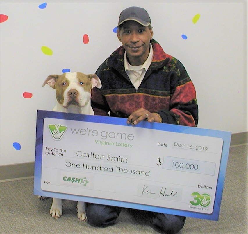 Image description: the prizewinner crouches on the ground holding his large check and his good boy Zeus sits perfectly beside him looking straight at camera.