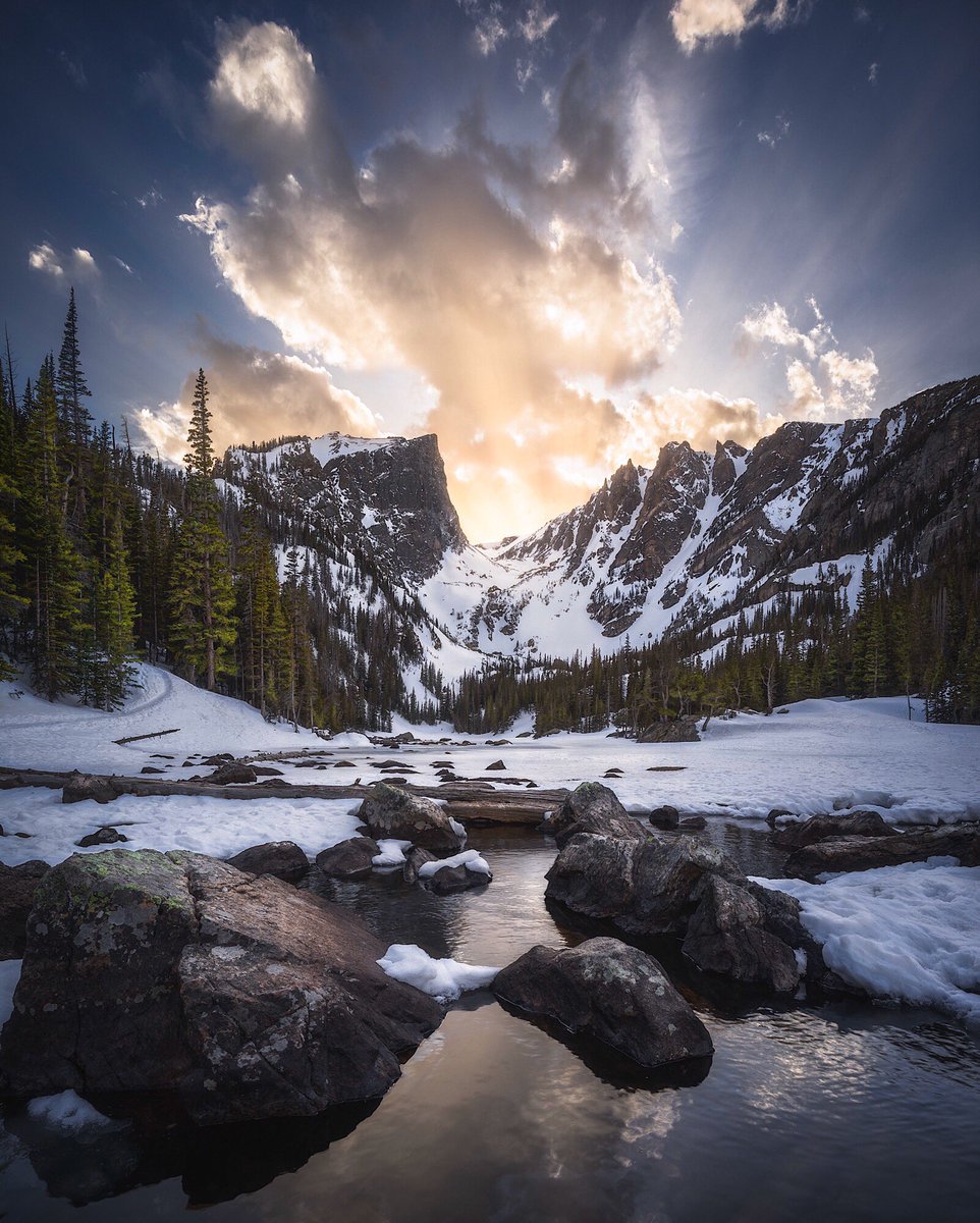 A mostly frozen lake sits at the base of a snow covered mountain under a sunset sky.