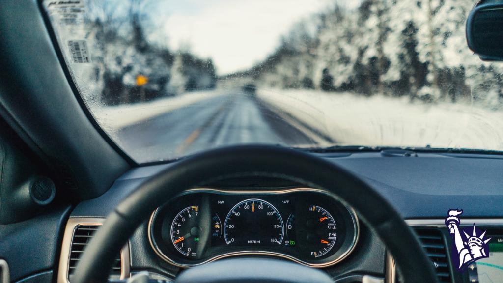 A photo taken from behind the steering wheel of a car. The car is driving down a snowy road lined with trees. 