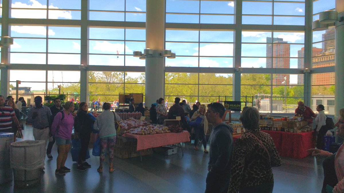 A crowd of people are seen shopping at a farmers market with tables of items inside the Whitehall Ferry Terminal.