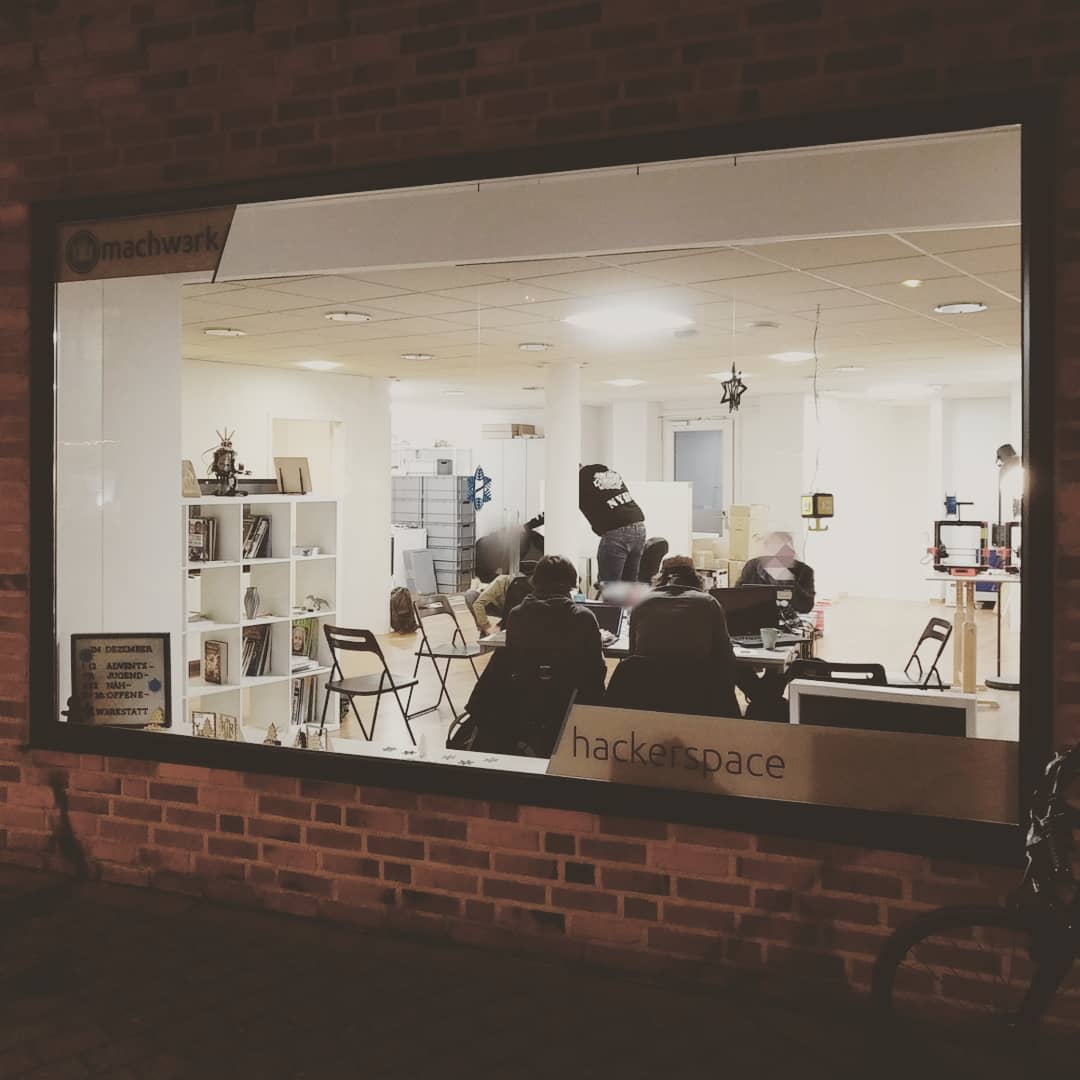 Von außen aus dem dunklen in ein hell beleuchtete Schaufenster des Machwerk fotografiert. Es sind Menschen an einem Gruppentisch und stehend im Hintergrund zu sehen. Ca. 6 Personen. Es wird u.a. an Laptops gearbeitet