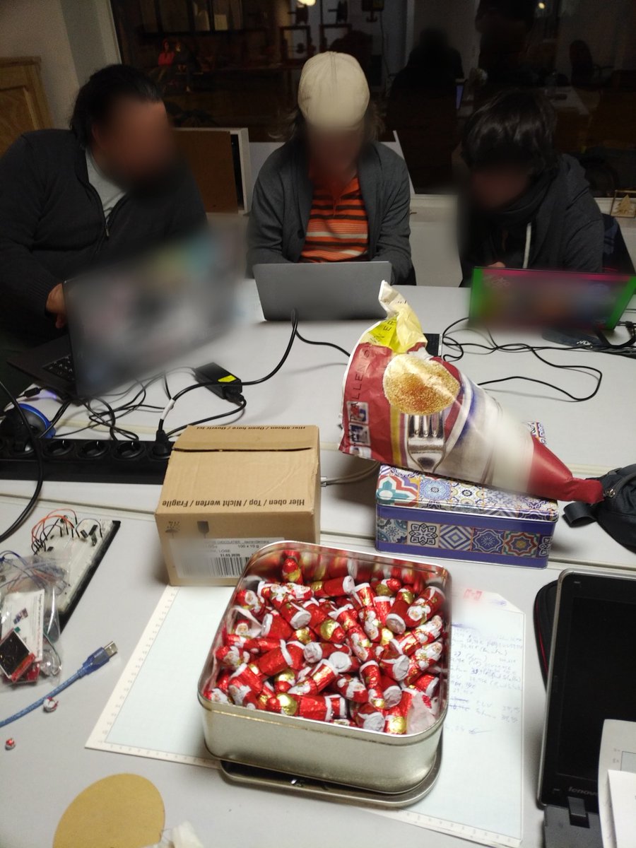 Drei Personen nebeneinander am eigenen Laptop. Auf dem Tisch Kabel, eine große Kiste voller Mini-Weihnachtsmänner und einer Papiertüte mit vermutlich Krapfen.