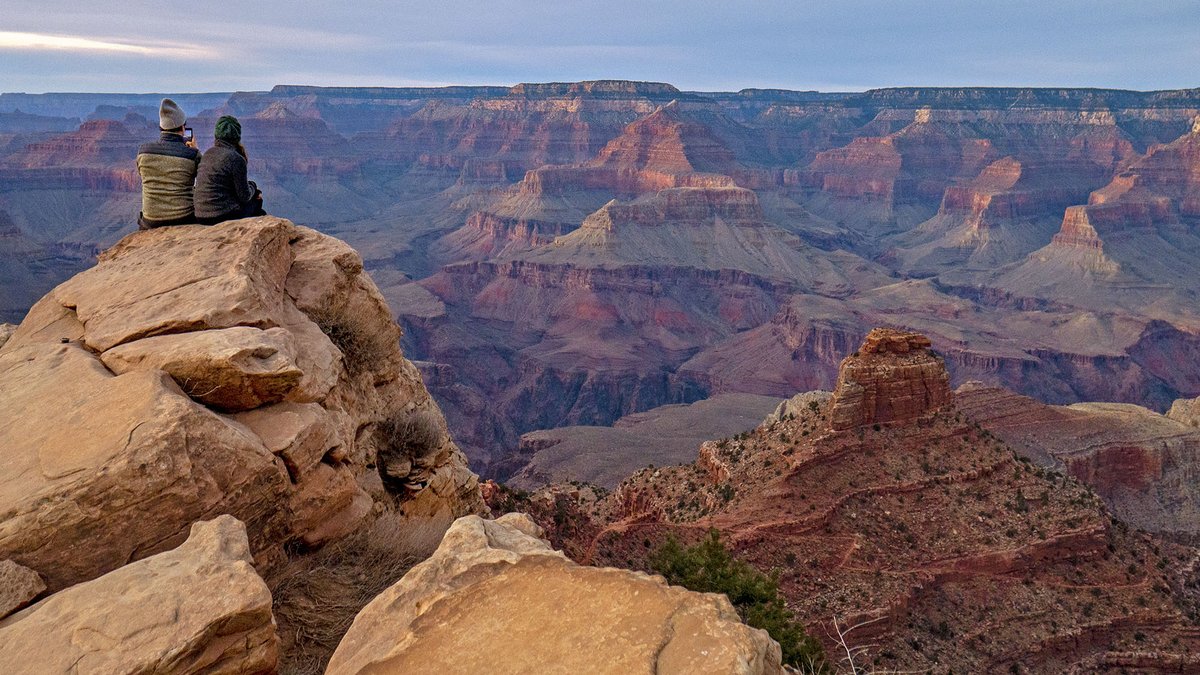 Description: two people sitting on a rock outcrop watching sunset inside a vast canyon of colorful peaks and cliffs. NPS/M.Quinn