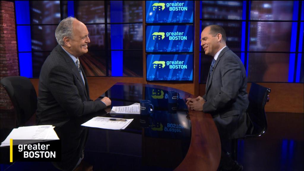 a smiling man sits behind an anchor desk as another man sits across from him speaking with his hands pressed together 