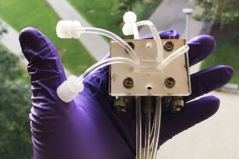A small metal box with many clear tubes protruding from it rests in the hand of a person wearing a purple latex glove.