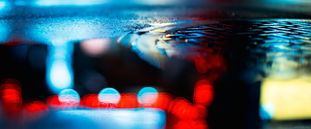 Photography by Liam Wong of Tokyo at night. Captured on a 50mm lens - the focus of this set of images is 'bokeh' and 'rain'. This image has the reflection of the Kabukicho gates neon sign and is a close up of a manhole. It is red, blue and a hint of yellow.