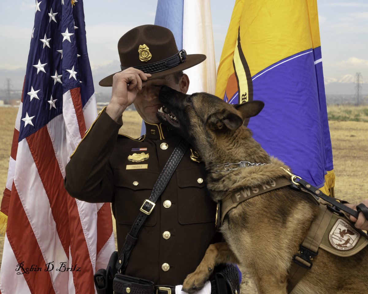 officer in full uniform holding brim of hat while german shepherd licks his face