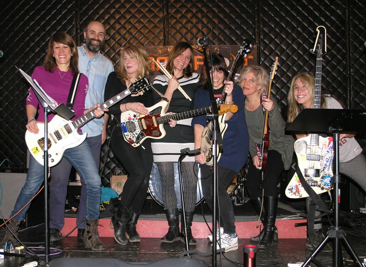 I'm standing in a line with Jen, Hilken, Freda, Jennifer, Tanya, and Gail in a rehearsal studio. Freda is holding up her drumsticks; the others are holding up their guitars. All are smiling.