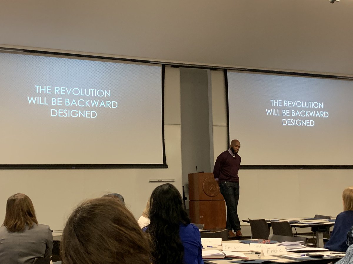 Dr. Dewsbury speaking on stage in front of a projection that reads “THE REVOLUTION WILL BE BACKWARD DESIGNED”