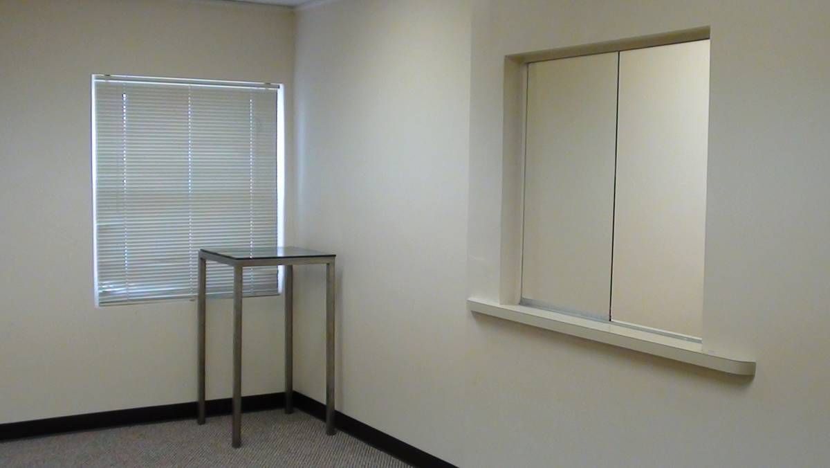 A photo of the corner of a room with beige walls. On the left, there is a table in front of a window covered with blinds. On the right, there is a receptionist's window with a sliding glass pane. The low-pile carpet is gray and the lighting is fluorescent.