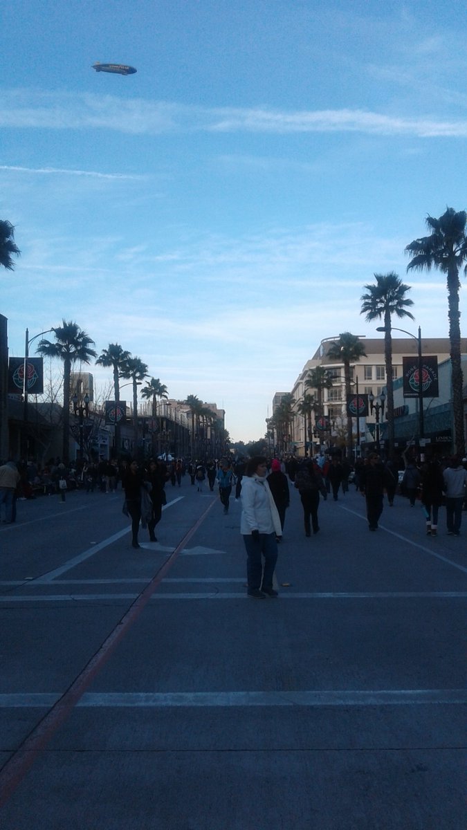 Palm trees line a human-populated street. A blimp floats overhead. 