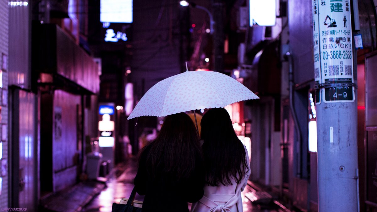 Photography by Liam Wong of Tokyo at night. A female couple share an umbrella through the rain. This image focuses on a closer shot of the couple.