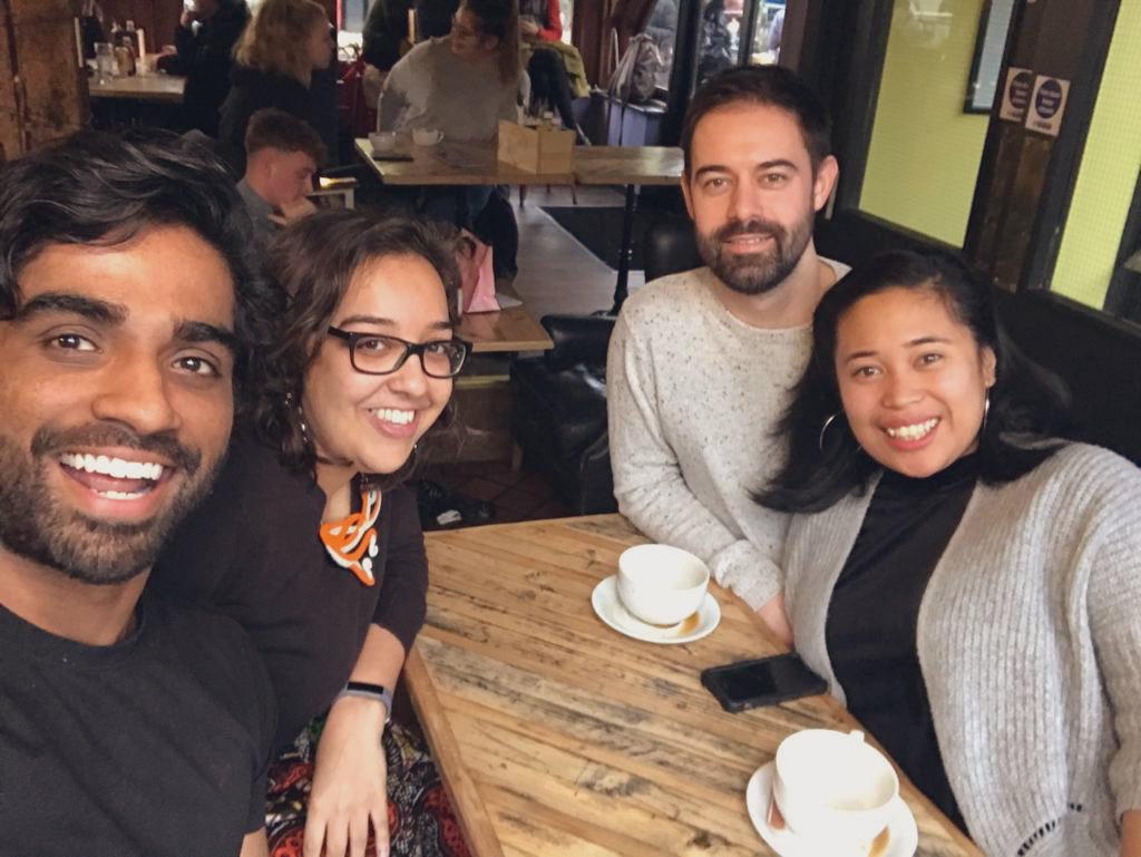 Four people take a selfie around a narrow wooden table with 2 empty coffee cups on it. To the left: South Asian male and female smile. To the right a southeast asian female and white male smile at the camera.