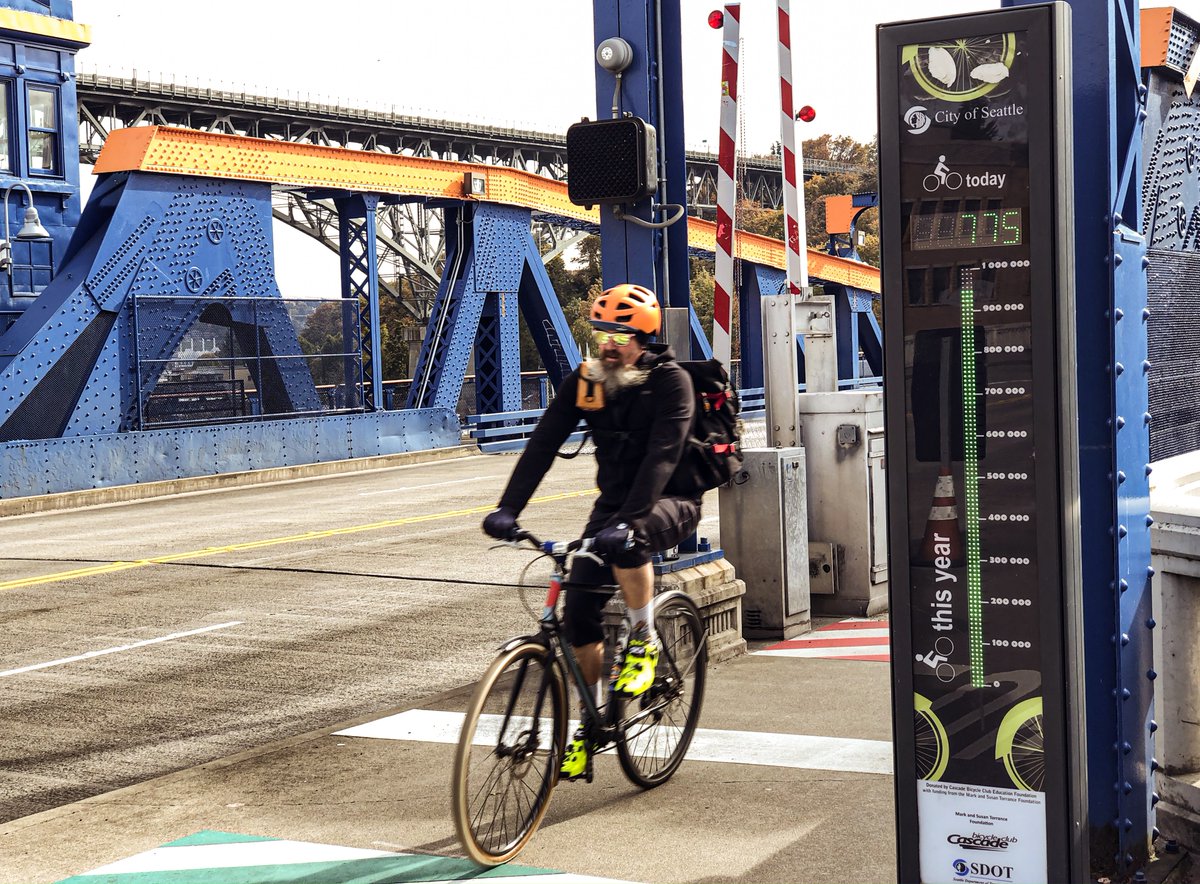 Photo of a person biking past the Fremont Bridge bike counter, a tall black structure with thermometer-like lights up the front tracking annual totals and a display of numbers counting the day's total.