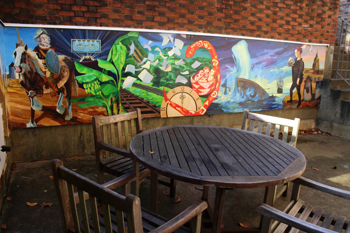 Nestled next to Tucker Hall is a small entryway with stairs leading down to a small courtyard. A table wooden table is placed at the center of the space, surrounded by four weathered chairs. On the wall is a large and vibrant mural.