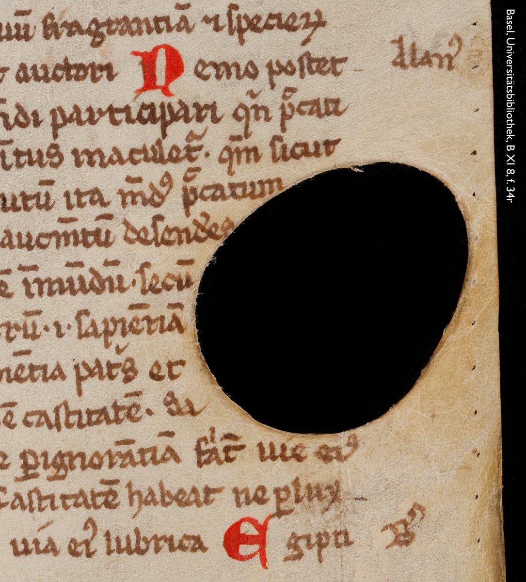 Photo of a part of a leaf from a medieval manuscript showing 13 lines of abbreviated Latin copied in brown ink & decorated with bright red majuscules. The parchment leaf is an offcut—a lower-quality scrap from the edge of the treated hide. Towards its edge is a large hole surrounded by areas of discolouration. Shelfmark: Basel, UB, B XI 8, f. 34r.
