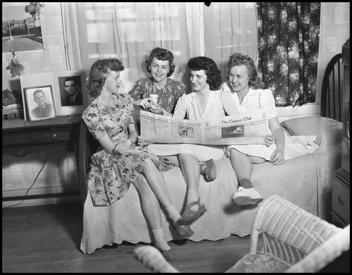 Black and white photo of four women sitting on a bed each looking at another, with one woman holding open a newspaper, "The Campus Chat."