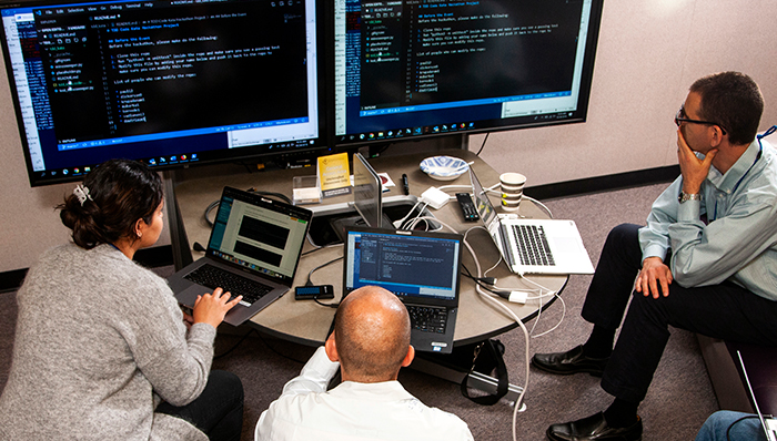 three people working around laptops and two large monitors
