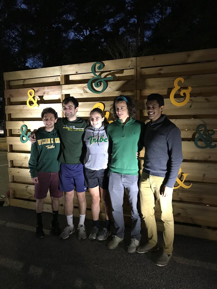 A group of students dressed in W&M attire smile and link arms in front of a wooden wall with floating green and gold ampersands at the 2019 Fire and Ice block party following the Charter Day ceremony.