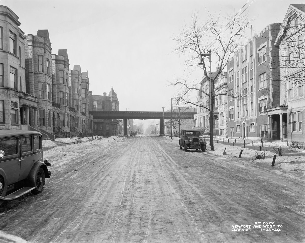 The view looking west from the center of Newport Avenue, a residential street. There are apartment buildings on each side made of stone or brick. A utility pole and a bare tree stand next to each other on the right side. The ground is covered with snow. There are three cars: one in the left foreground, one in the right background, and one in the far left distance. In the center background are elevated train tracks. 