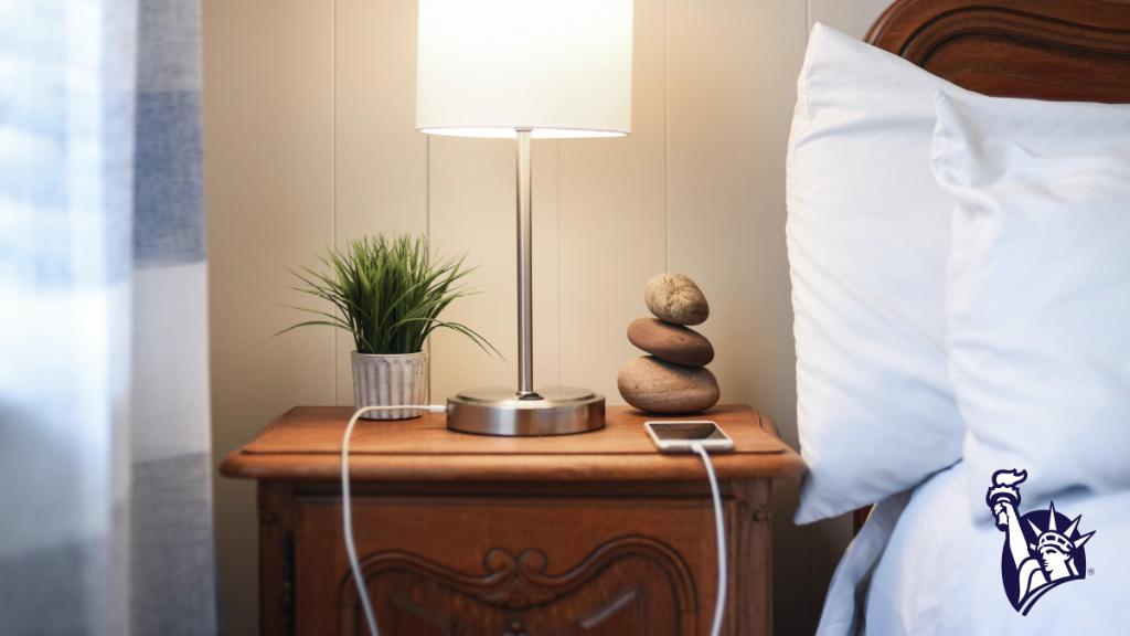 A lamp is turned out on a bedside table that also includes a small plant, rock sculpture, and a charging cell phone.