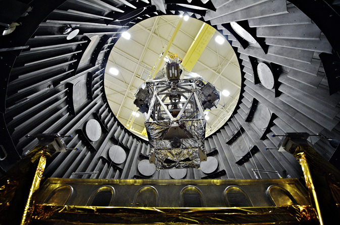 A spacecraft gets lowered into a large metal thermal vaccuum chamber. 