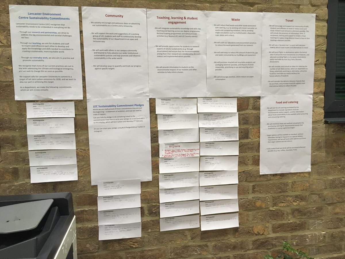 Pieces of A4 paper stuck to a brick wall over an industrial printer. Stuck under the A4 pages are strips of paper on which people have written their pledges