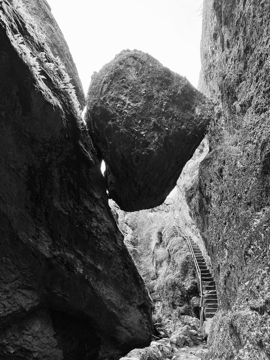 Black and white photo of a huge boulder wedged between two vertical rock walls. A stone stairway passes under the boulder and up behind it.