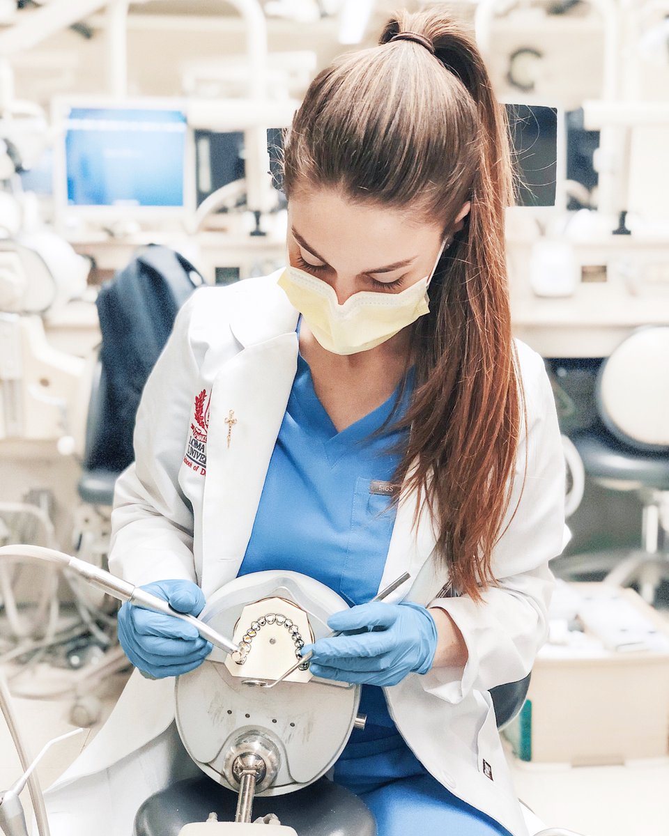 Charlotte wearing scrubs, a lab coat, and gloves holding dental tools. She is sitting behind a model of human teeth, using her tools on the model.