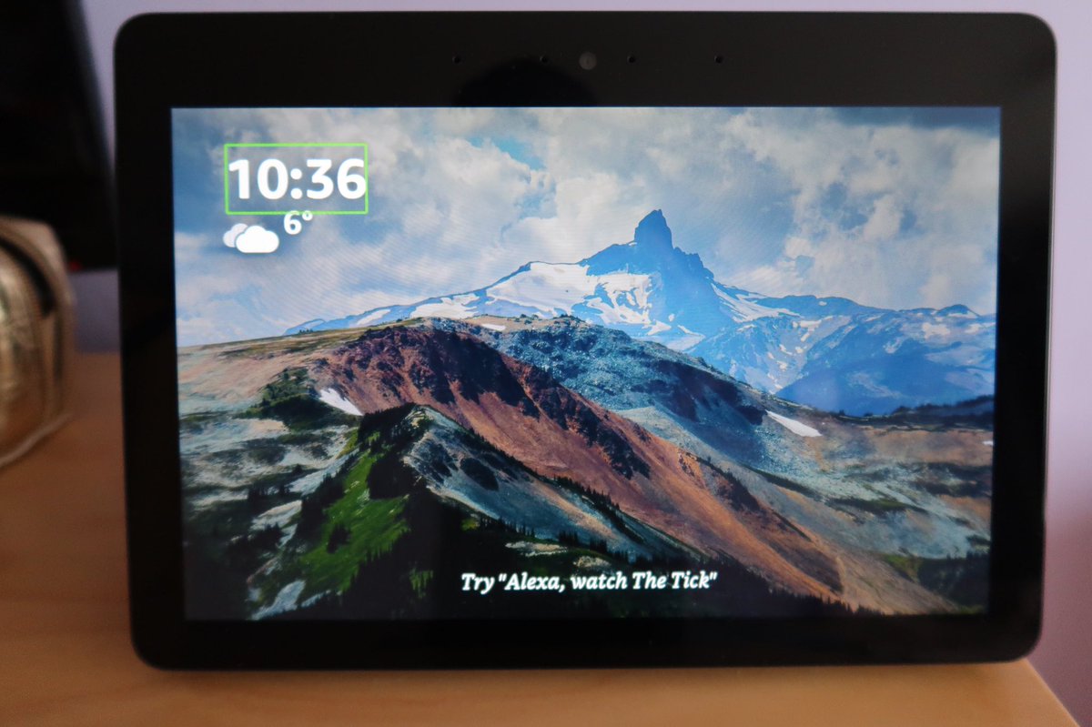 A photo of an Amazon Echo Show which has a background picture of mountains and clouds on the screen