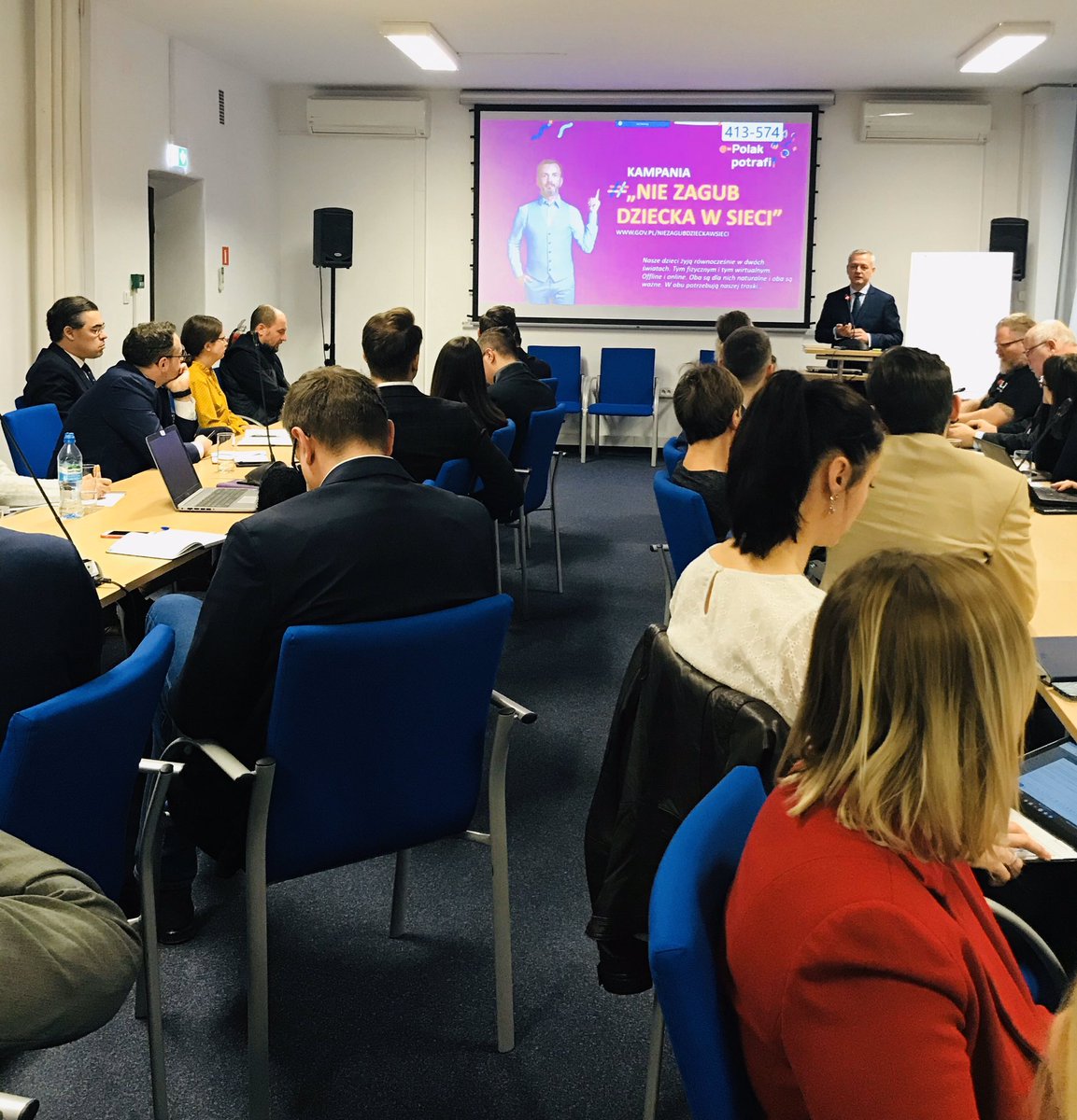 Minister Zagórski stoi na tle ekranu, na którym wyświetlana jest prezentacja. Przed nim siedzący członkowie grupy.