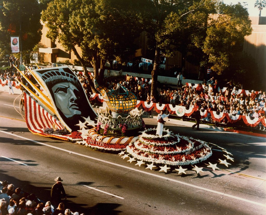 Photo of the original torch on display during the Tournament of Roses Parade. Three part float with original torch in the middle, face of the statue on left, and a woman wearing a white dress holding flowers on right.