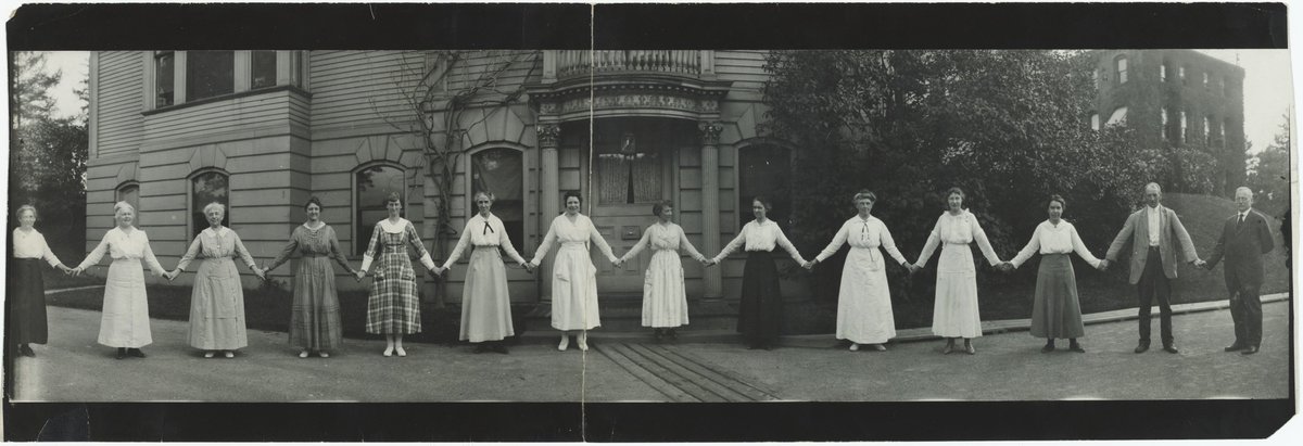 Black and white photo of 14 people, mostly women, standing about an arm's length apart, holding hands, in front of a building with Corinthian columns. The women wear long skirts. 