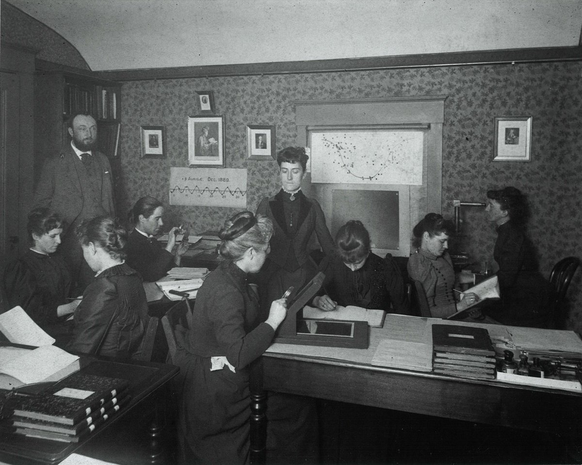 In a small office space, about eight women work at desks, books open and pens in hand. They look at their work, not the camera. A man in corner looks at the scene. 
