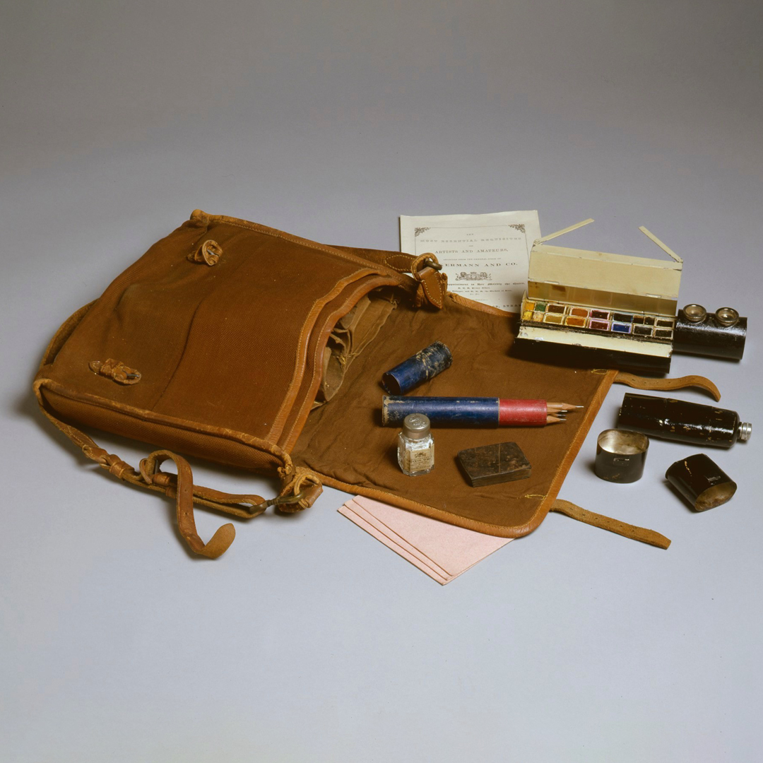 A leather satchel with various art tools and supplies laid out, including watercolours and pencils.