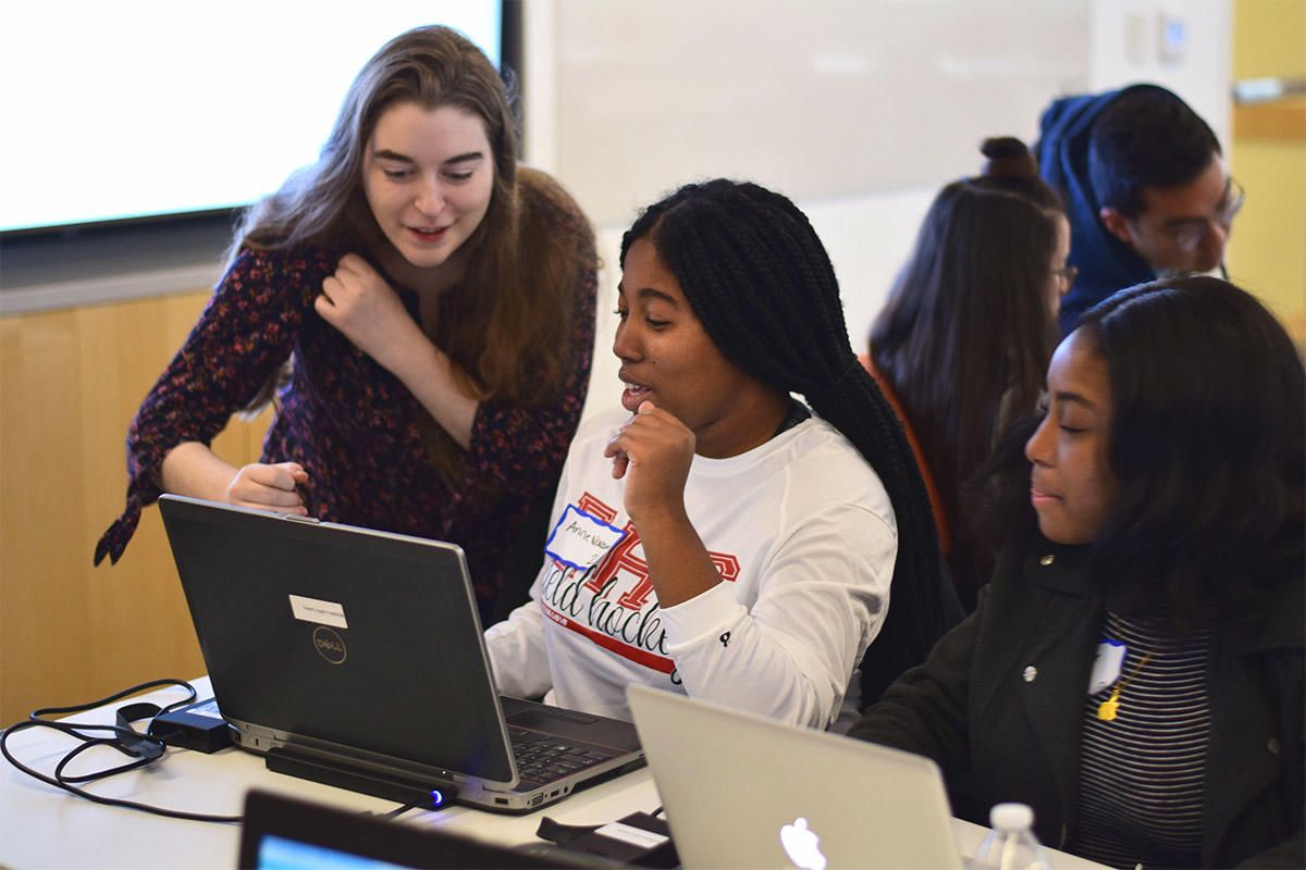 Students work on laptops while a woman assists them