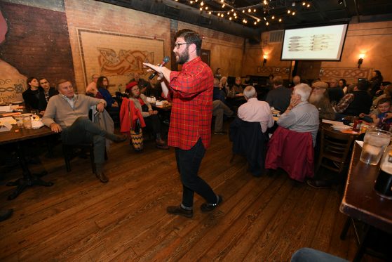A man stands in the middle of a pub speaking into a microphone. People sitting at tables around him are listening. 