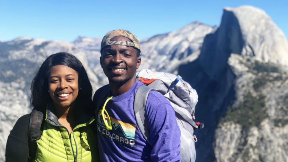 A young man and woman smile at the camera while standing in front of a gorgeous mountain landscape.