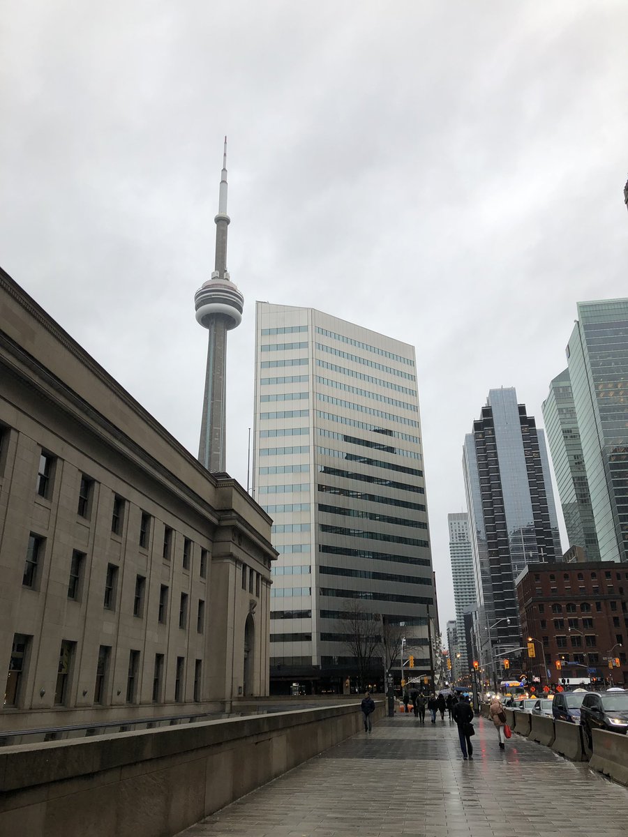 A view of some downtown Toronto buildings and the CN Tower against a cloudy grey sky. The sidewalk is wet and shining and some people are walking around without umbrellas out.