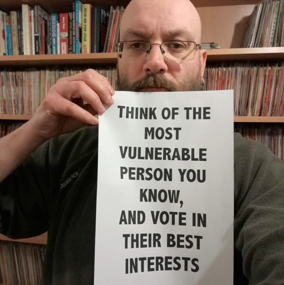 A man holding a piece of paper that says "Think of the most vulnerable person you know, and vote in their best interests."