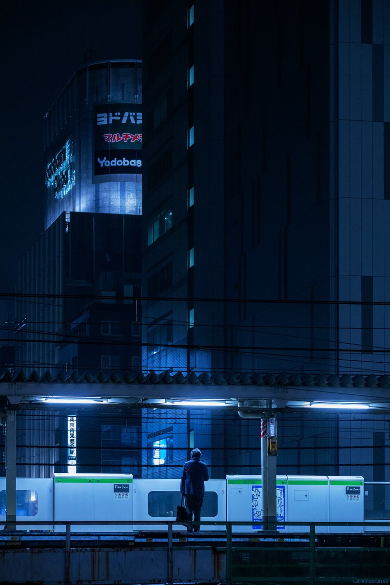 Photography by Liam Wong of Tokyo at night. A businessman waits on a platform by himself. The image is vertical and shows off the sheer scale of Tokyo's architecture. He is lit by three halogen lights overhead.