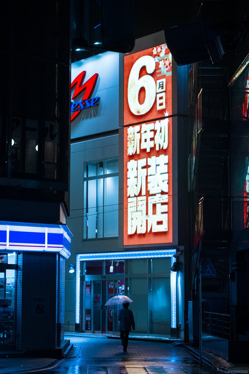 Photography by Liam Wong of Tokyo at night. A woman holding an umbrella walking down an alleyway. It is a vertical image and a large neon sign is above her in red. To the left is a konbini / Lawson convenience store. 