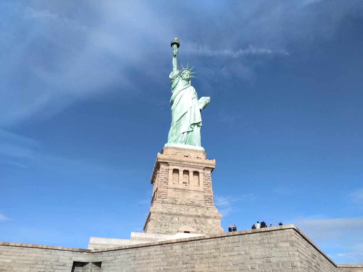 A photo of the Statue of Liberty with blue sky and clouds in background and a few visitors on top of Fort Wood.