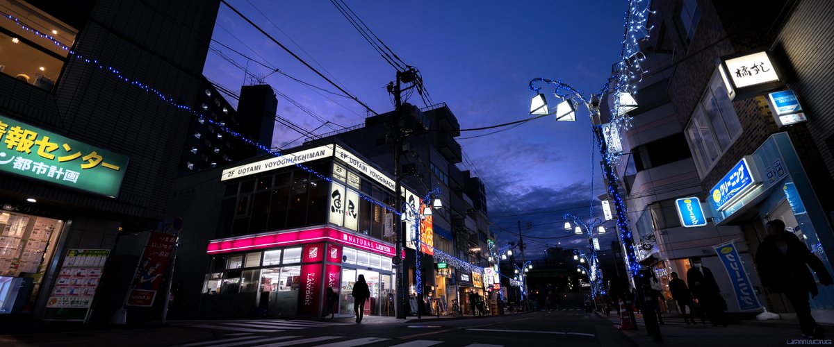 Photography by Liam Wong of Tokyo during the evening. A wide street shot - on the left side the silhouette of a woman and her child walking in front of a convenience store - the shop front is pink in color. The sky is lavender and has light clouds. Wires cut through the sky.