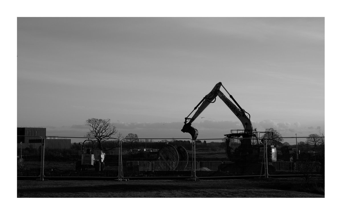 Black and white. A digger's arm and a bare tree are the only things seen above the construction fence, all else is level