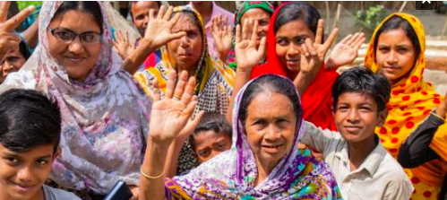 A group of women and children dressed in colourful garb face the camera raising one hand in the air, signifying their collective unity.