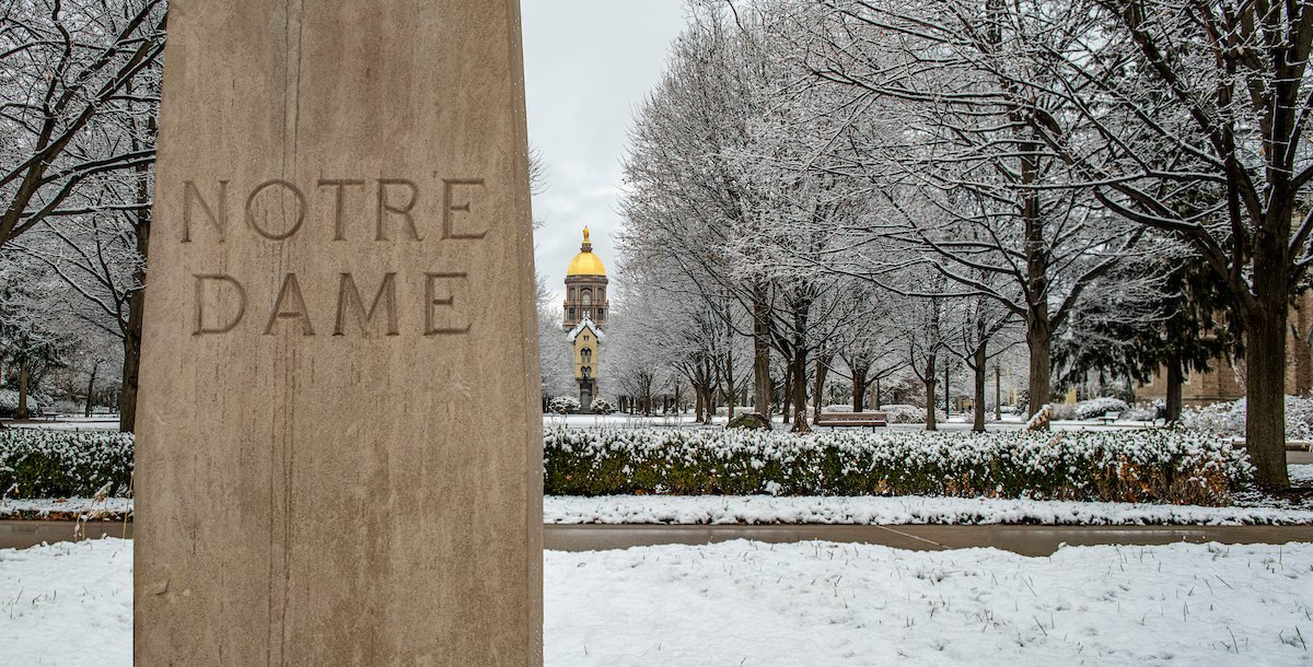 A pillar reading Notre Dame in front of a snowy scene of the Golden Dome of the University of Notre Dame