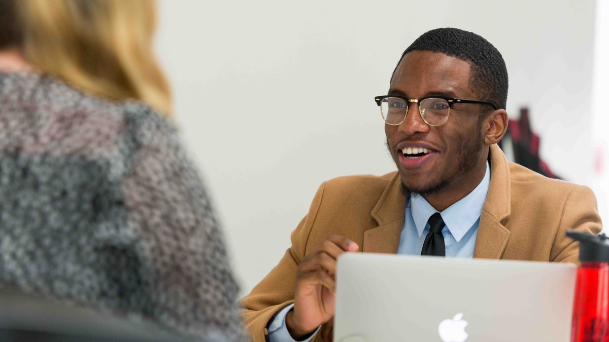 Male student in business attire speaking a colleague with a laptop.