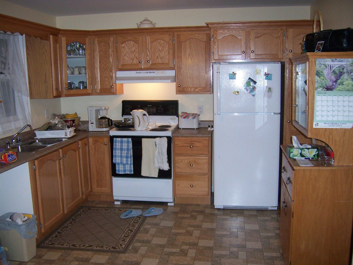 A flash photo of a cluttered residential kitchen with wood cabinets, a linoleum countertop, white appliances and a patterned brown linoleum tile floor. There is a boombox on a cabinet to the right. On the counter and stovetop, there are appliances, a drying rack full of dishes and a water pitcher. There is a pair of slippers on the floor near a small brown area rug. There is a trashcan on the left.