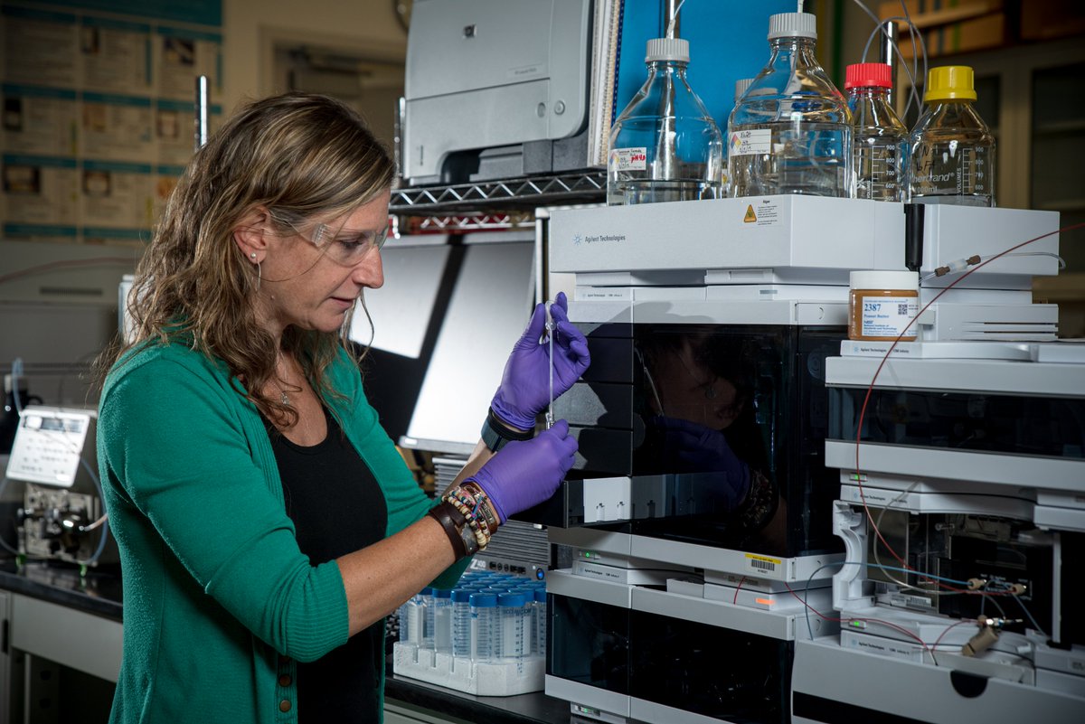 Melissa wearing goggles and gloves, pipetting liquid into a vial. She is standing in front of scientific instruments. 